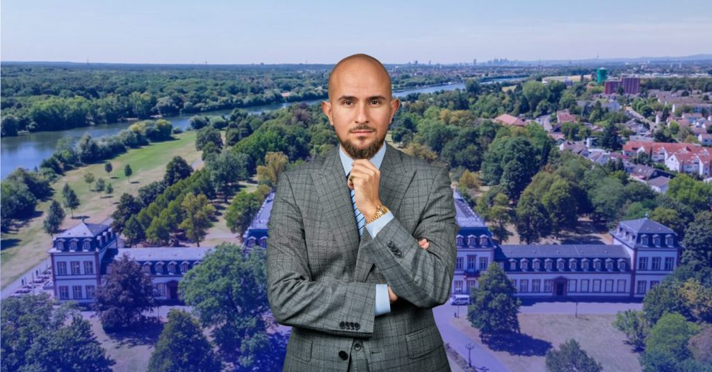 Ein elegant gekleideter Mann in einem grauen Anzug mit Krawatte steht vor einer malerischen Landschaft mit einem historischen Gebäude und weitläufigem Grün. Im Hintergrund sind ein Fluss, Bäume und eine Wohngegend zu sehen. Das Bild vermittelt einen professionellen Eindruck und symbolisiert Expertise im Bereich Immobilien kaufen.