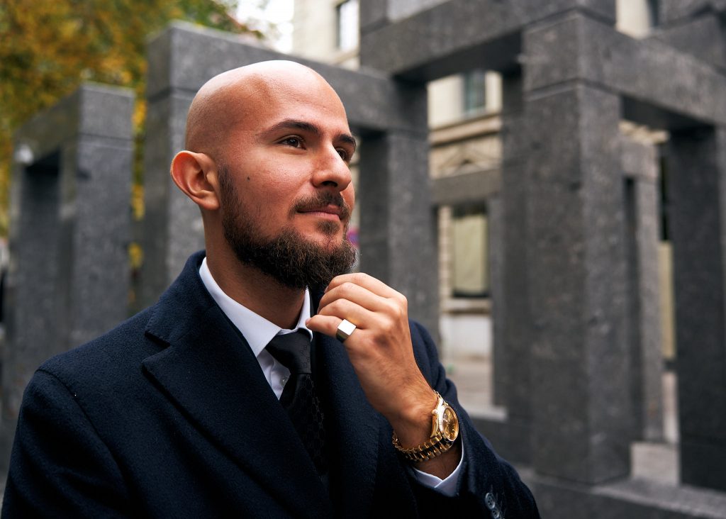 Alexander Tsasakos, stilvoll gekleidet in einem dunklen Mantel und Anzug, steht vor einer modernen Architekturkulisse. Er wirkt nachdenklich und selbstbewusst, während er seine Krawatte richtet. Das Bild vermittelt Professionalität und Expertise im Bereich Investieren in Immobilien.