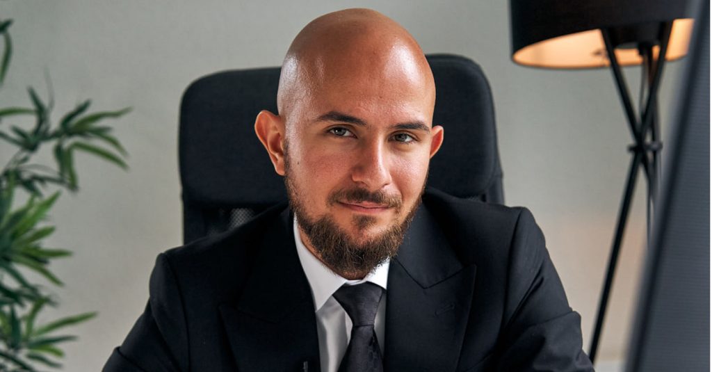 Ein professionell gekleideter Mann in einem schwarzen Anzug und Krawatte sitzt an einem Schreibtisch. Im Hintergrund sind eine Zimmerpflanze und eine moderne Stehlampe zu sehen. Der Mann wirkt selbstbewusst und freundlich, was Expertise im Bereich Immobilien und Kapitalanlagen symbolisiert.